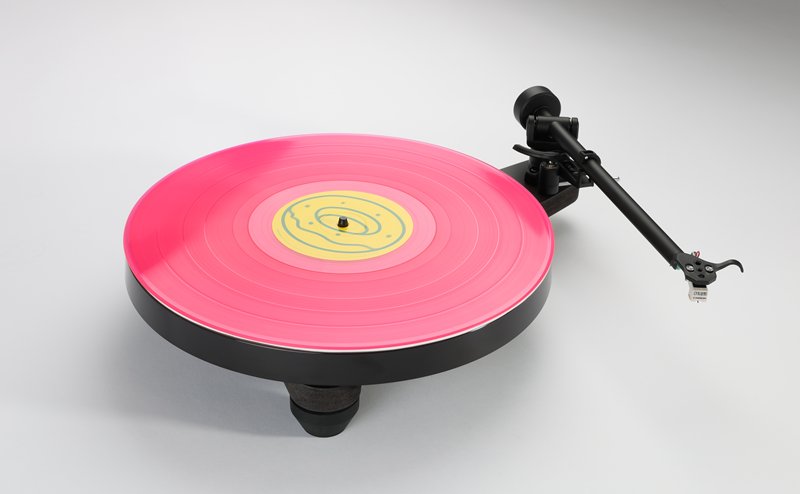 The Rega Replay in matt black, three-quarter view from above, a pink record on the platter and the RB110 tonearm on its rest.